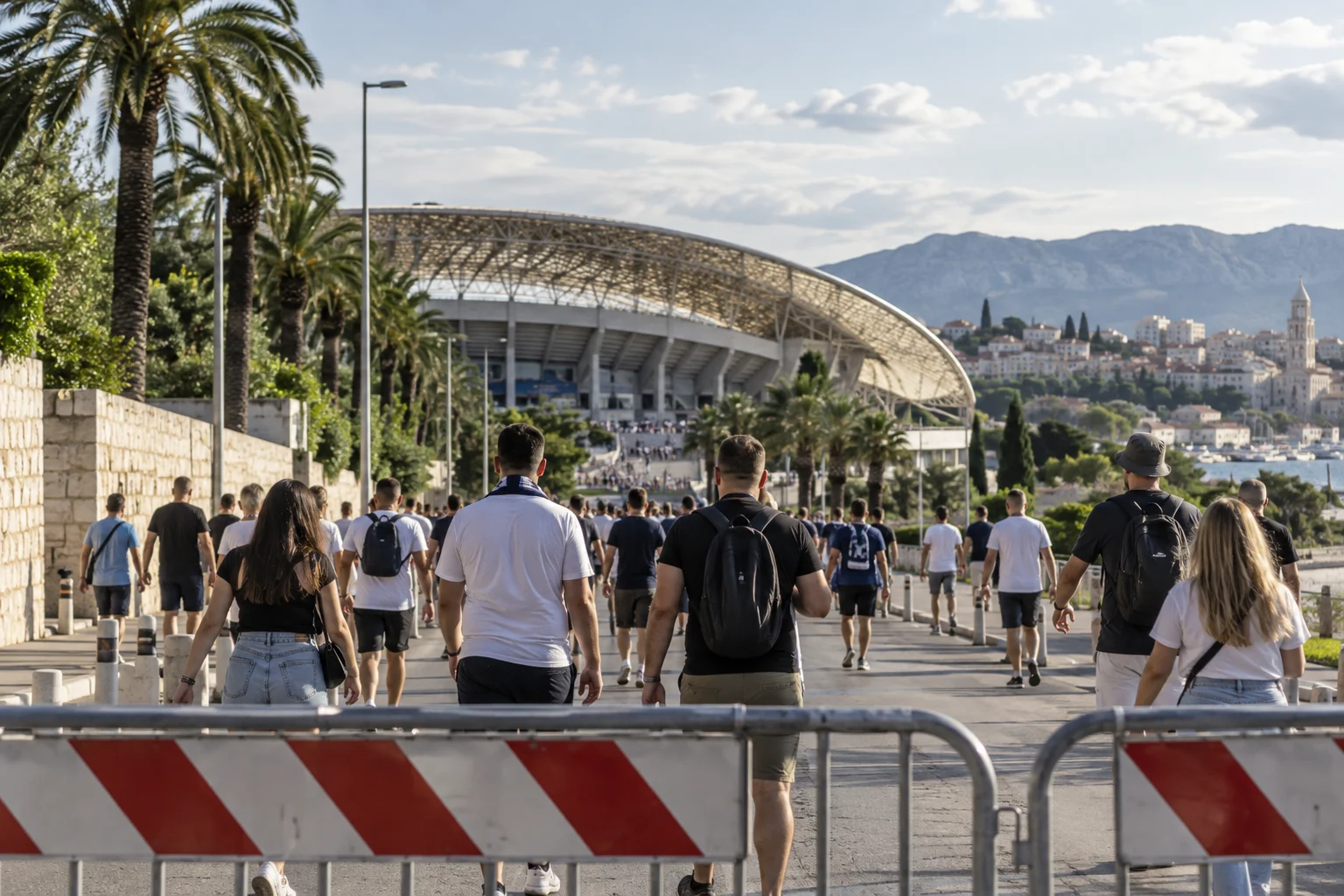 Besucher gehen zu einem mediterranen Stadion, im Vordergrund steht eine vorübergehende Straßensperre